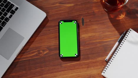 smartphone with green screen top view. mobile phone touchpad with chroma key close-up, laptop computer and tea cup on table.