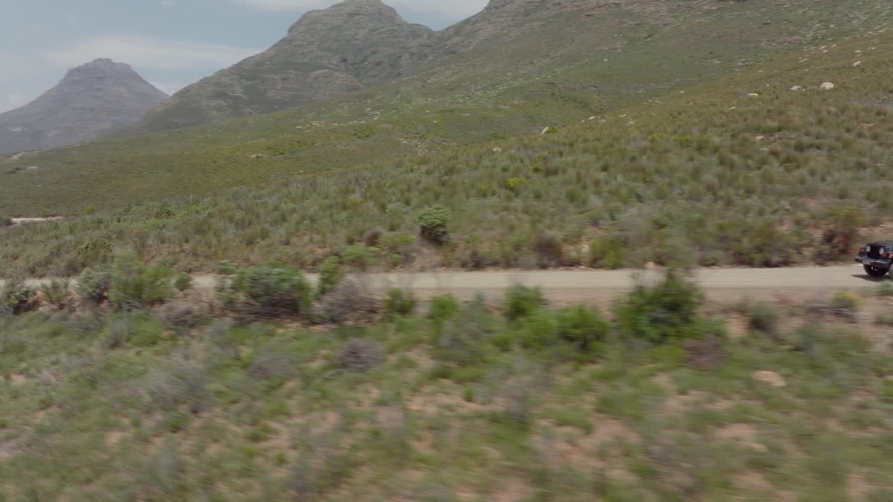 drone vuela rápidamente al lado de una carretera y paisaje rocoso en el área salvaje de cederberg, sudáfrica con una vista de un vehículo todoterreno conduciendo en una carretera - se pueden ver las montañas y la cola
