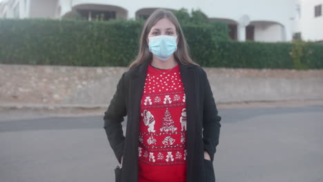 young adult wearing a christmas sweater putting on a surgical mask in the street and looking at the camera