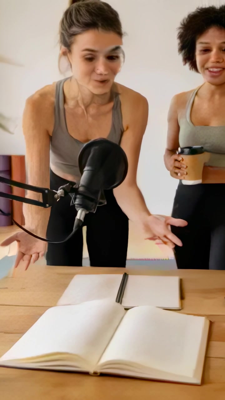 Video vertical: Pasando páginas, dos mujeres con camisetas grises inclinadas en una mesa, encontrando páginas sueltas cerca de un micrófono.
