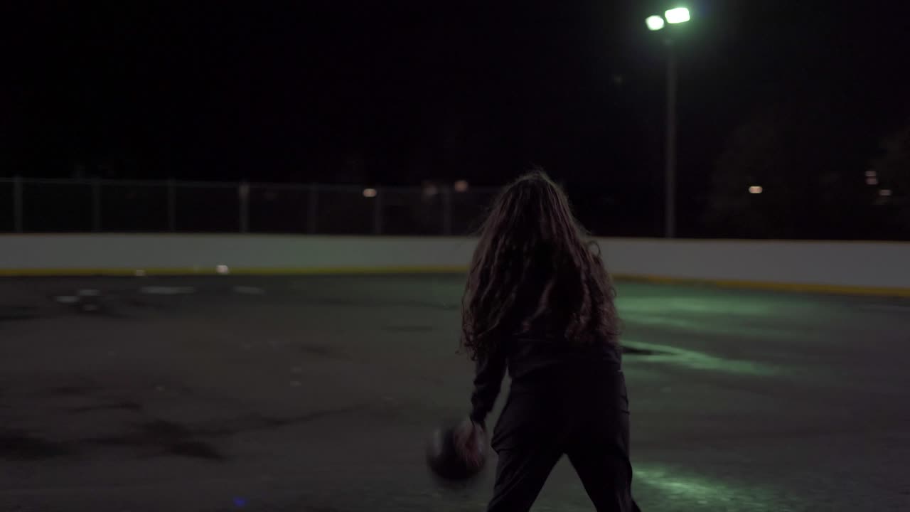 Teenage girl dribbles ball then shoots it at net at an outdoor court at night