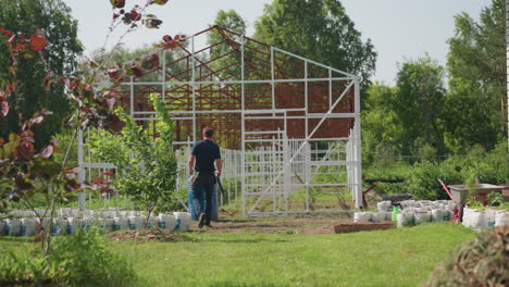back view of man walking toward farm carrying rifle, grow bags and white metal frame structure ahead, trees beyond fence, summer rural scene with grass underfoot and soft sunlight on plants