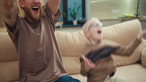 A-happy-blond-man-with-a-beard-and-glasses,-together-with-his-little-albino-son,-are-rooting-for-their-favorite-football-team-and-watching-the-match.-The-little-albino-boy-is-holding-a-soccer-ball-in-his-hands-and-then-throwing-it-up.-After-the-happiness-of-Victory