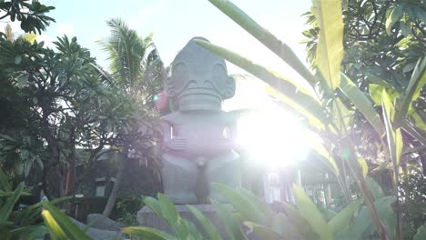 large carved tiki statue in the tropical islands of french polynesia with palms and a beautiful light flare