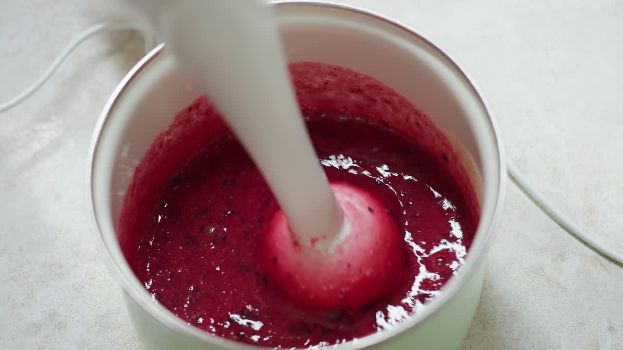 Close up of person blending thick currant berry mixture using electric hand blender in kitchen pot, showing swirling movement and texture of deep red puree during food preparation on light countertop