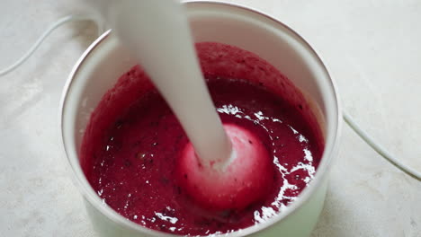 close up of person blending thick currant berry mixture using electric hand blender in kitchen pot, showing swirling movement and texture of deep red puree during food preparation on light countertop