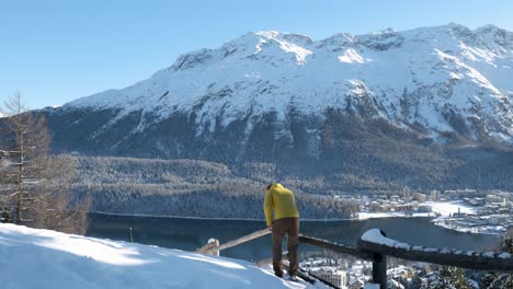 joven caucásico con una chaqueta amarilla y un gorro disfrutando de la vista de un lago, una ciudad y montañas en un soleado día de invierno en st.