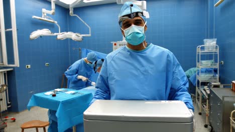Portrait-of-surgeon-holding-ice-box-while-colleagues-performing-operation