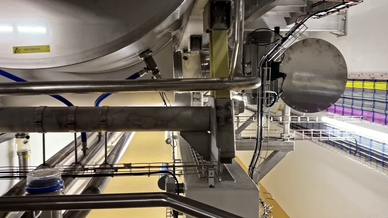 Panning shot inside modern food processing facility showing extensive multicolored cable trays piping stainless steel tanks hoppers and hygienic conveyor systems under bright lighting