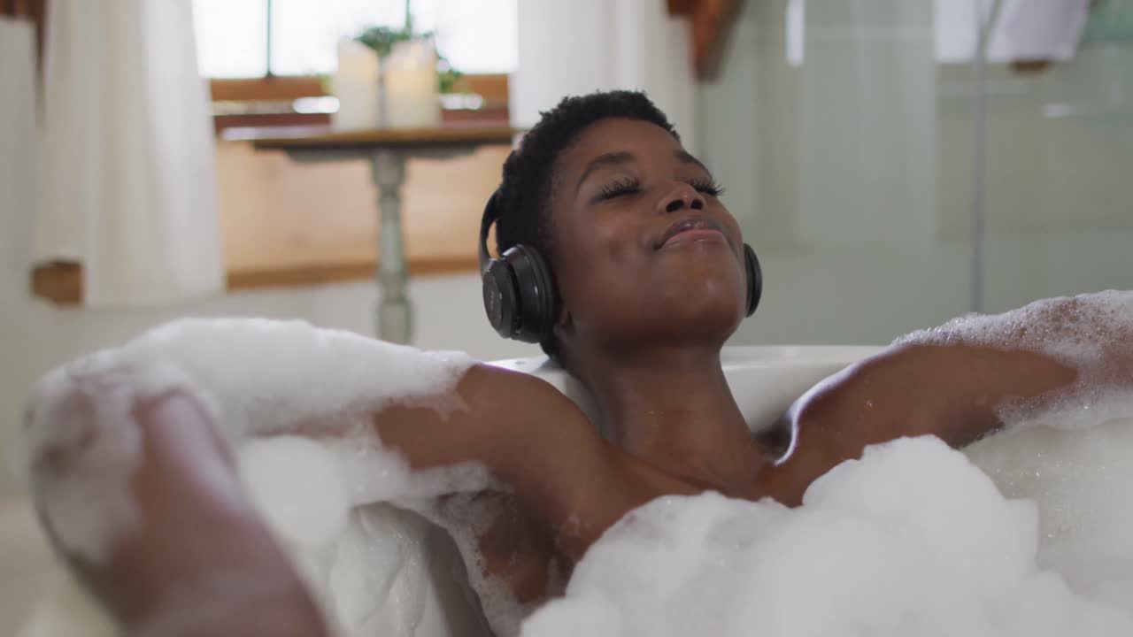 미소 짓는 아프리카계 미국인 매력적인 여성은 헤드폰을 착용하고 욕실에서 거품 욕조에서 휴식을 취하고 있습니다.