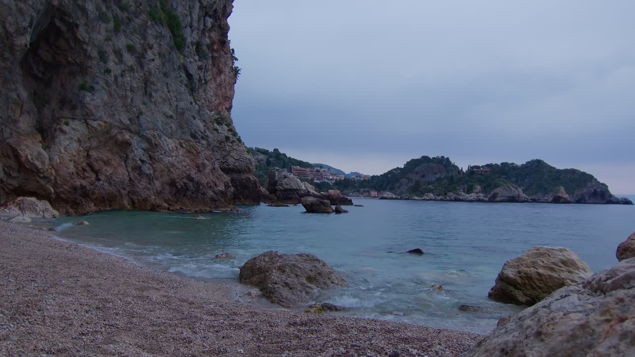 Dolci onde che lambiscono una spiaggia con grotta con l'Isola Bella incorniciata in lontananza a Taormina, Sicilia, Italia, cinematografico e tranquillo