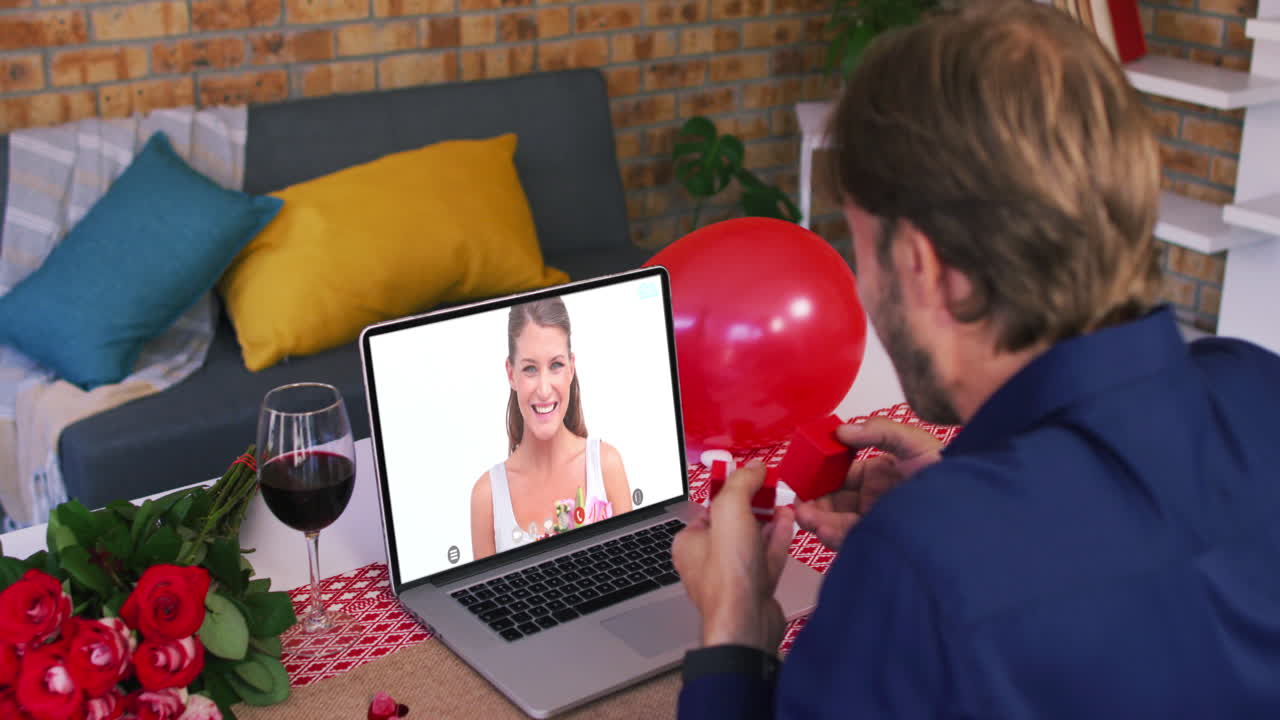 Casal caucasiano em videochamada de Dia dos Namorados, homem mostrando anel para mulher sorrindo na tela do laptop.