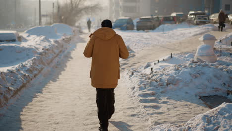 back view city dweller wearing insulated winter coat walking on snow covered urban sidewalk flanked by parked cars and modern buildings while exhaling misty breath into crisp frosty morning light