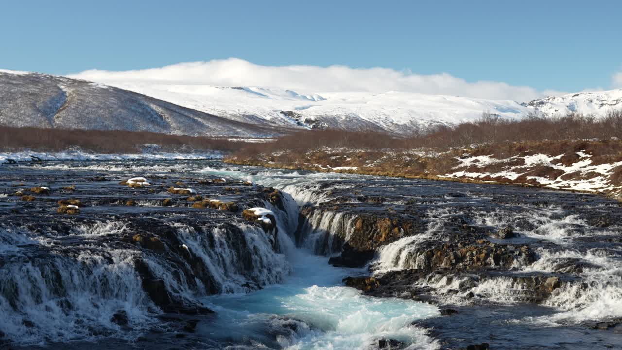 Wide static view of Brúarfoss waterfall in Laugarvatn, Haukadalur, Iceland, with vivid blue glacial water flowing through dark volcanic rocks under a snowy mountain landscape.