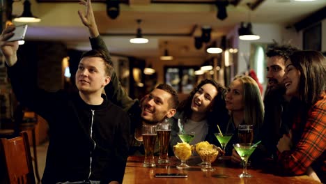 young blogger is recording video with smartphone sitting at table in bar with his friends. young people are posing, waving hands and laughing. blogging for instagram concept.