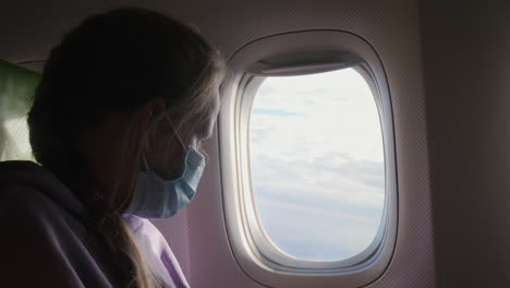 una niña de diez años con una máscara de protección mira a través de la ventana del avión. vacaciones durante la pandemia de coronavirus