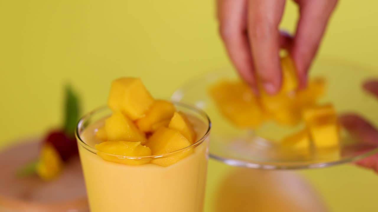 Hand adds mango cubes to smoothie in glass, bright yellow background, soft natural lighting