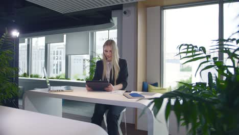 stylish business woman with tablet in office. elegant woman in suit sitting at table with laptop and surfing tablet in modern light office with big windows