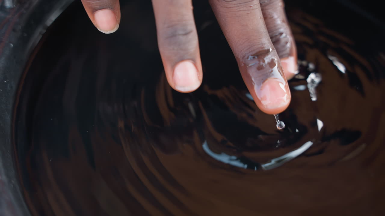 Dita di apprendista che mescolano mordente scuro per legno, con un movimento a spirale che crea increspature lucide. Mani che testano la miscela di colore e vernice. Pratica artigianale concentrata in officina, tecnica e studio attenti.