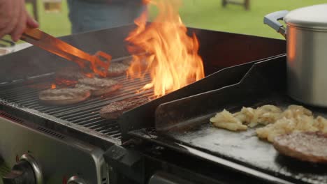 Mittlere-Aufnahme-Einer-Männerhand,-Die-Tagsüber-Burger-In-Den-Flammen-Eines-Grills-Im-Freien-Wendet