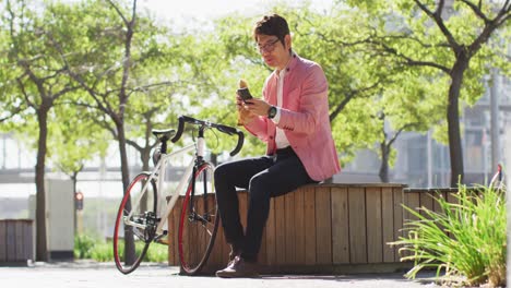 Asiatischer-Mann,-Der-Einen-Snack-Isst-Und-Sein-Smartphone-Benutzt,-Während-Er-Im-Park-Sitzt