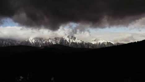 moody aerial drone shot of snow covered mountain range with silhouette mountain in foreground and moody grey clouds