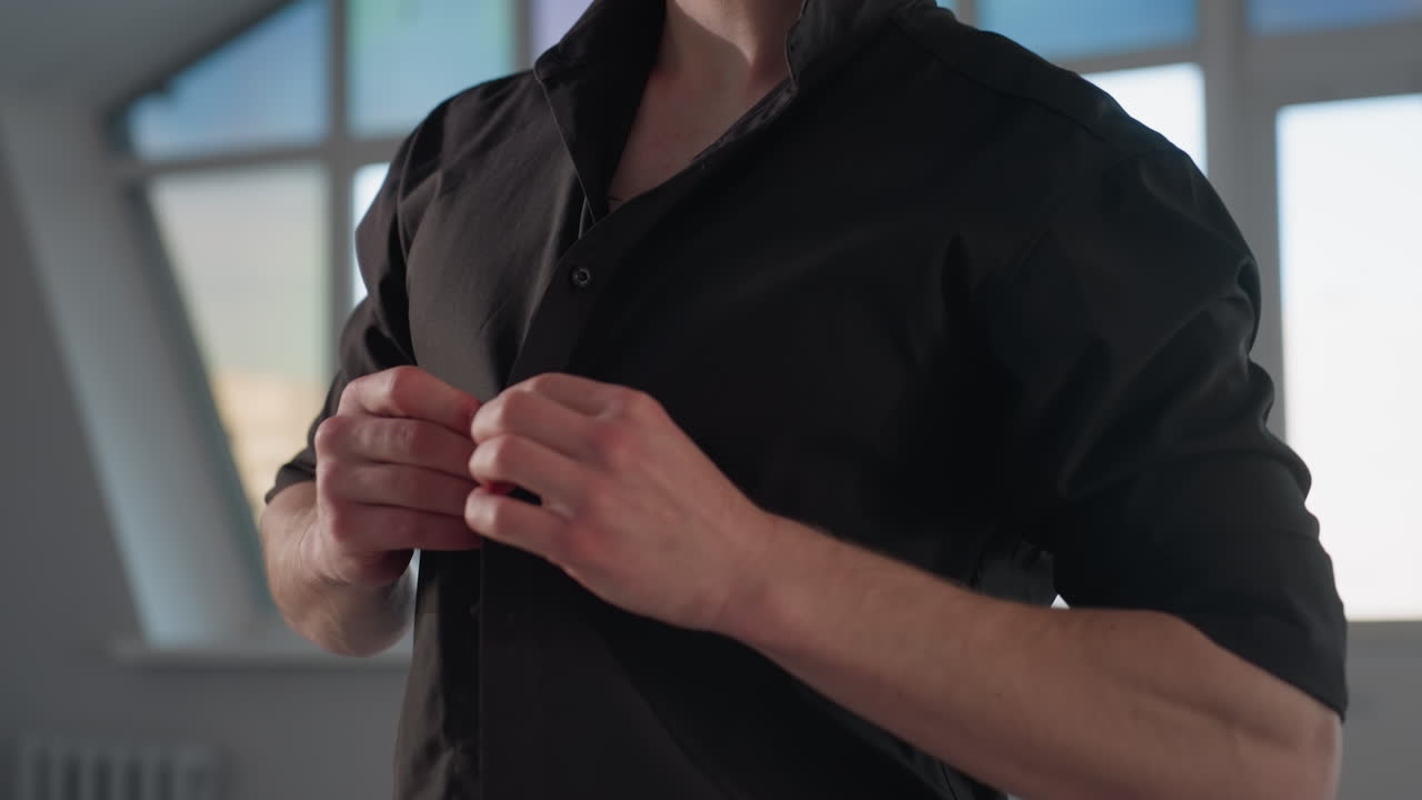 focused young man buttoning black shirt in front of bright loft window subtle neck tattoo visible contemplative gaze modern interior setting soft natural light urban backdrop mirror reflection