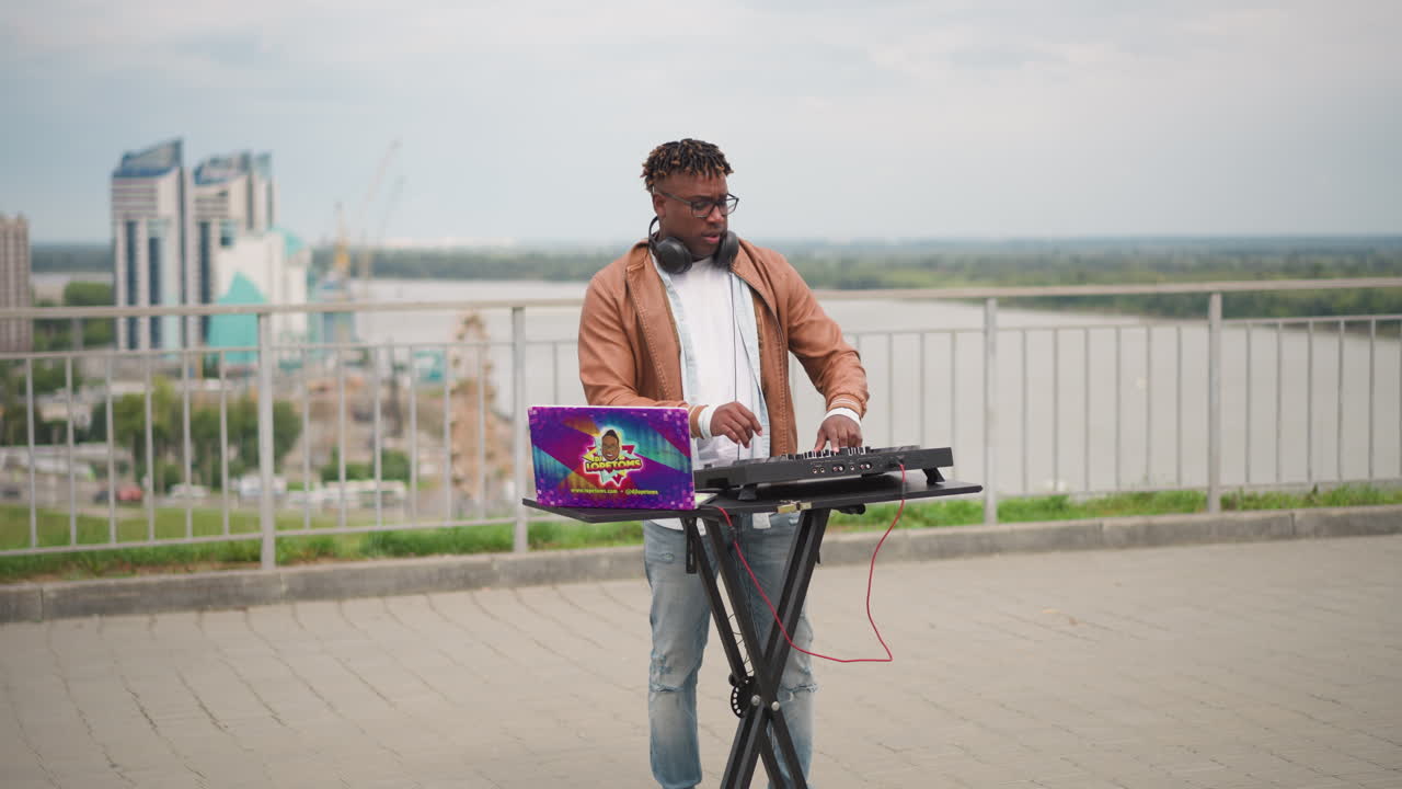 DJ on building rooftop blending melodies amid city panorama during daylight, Rooftop DJ wearing headphones and jacket crafting relaxing beats with panoramic city and river views during daytime