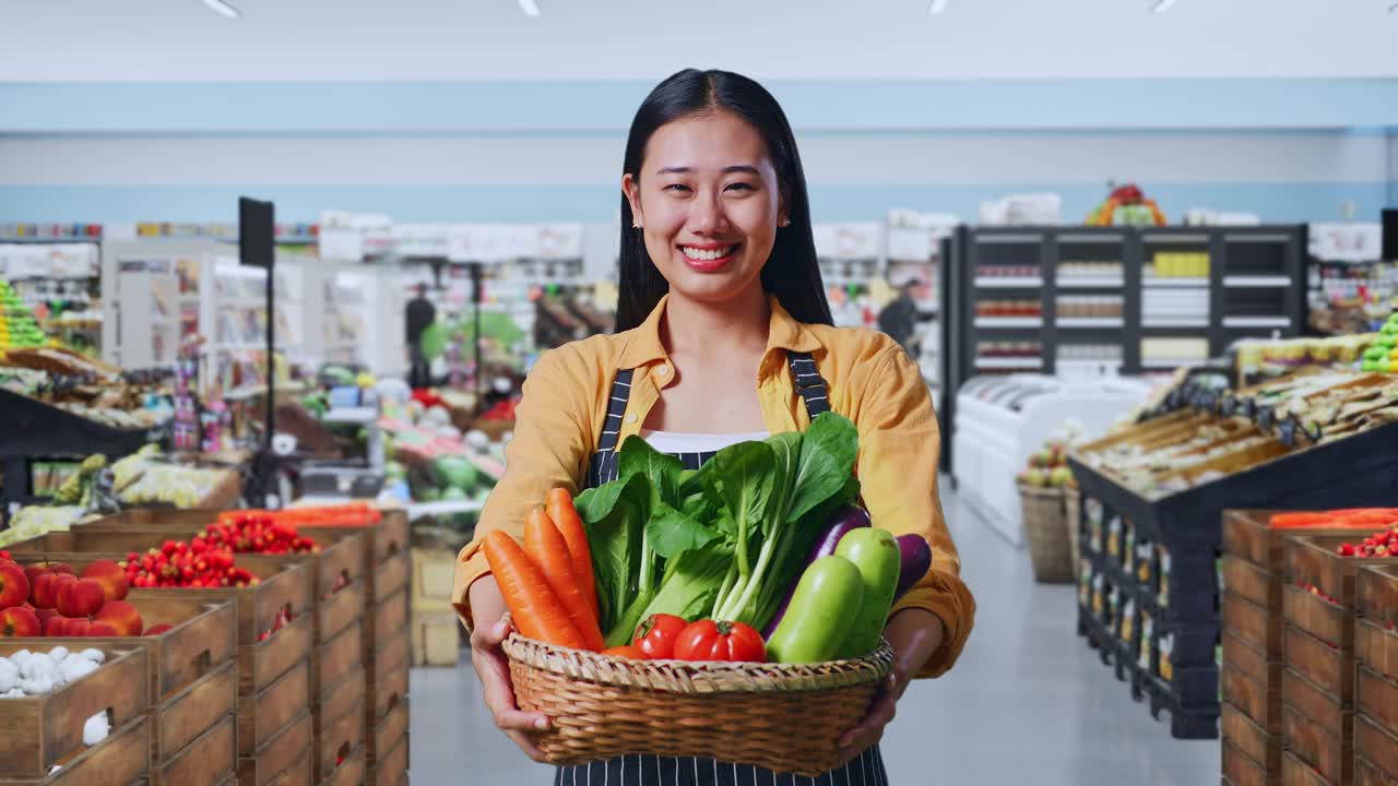 Agricultora asiática sonriendo amigablemente y entregando una cesta con verduras en una tienda de comestibles