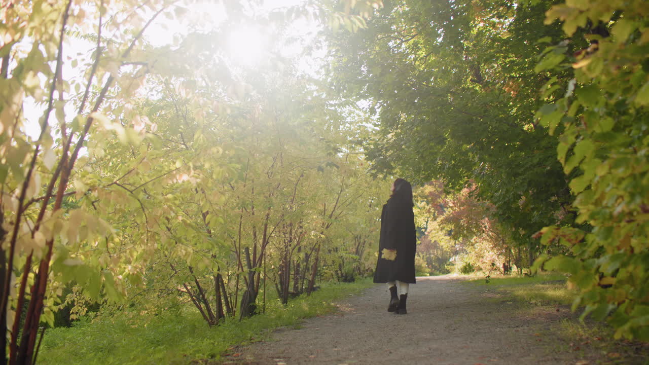 Long shot of autumn lover twirling under sunlit arboretum canopy, coat flowing as she turns along leafy path, golden trees framing joyful spin, quiet weekend wander through green tunnel
