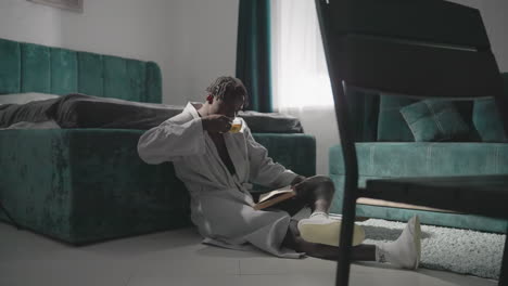 Man in white robe sitting on rug, reading book while holding tea cup. Bright morning light shines on modern bedroom with green furniture, creating relaxing and peaceful atmosphere for self-care