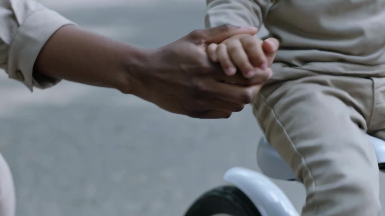 Reaching parent holding child's hand on park path, guiding bike teaching child to ride, wearing tan