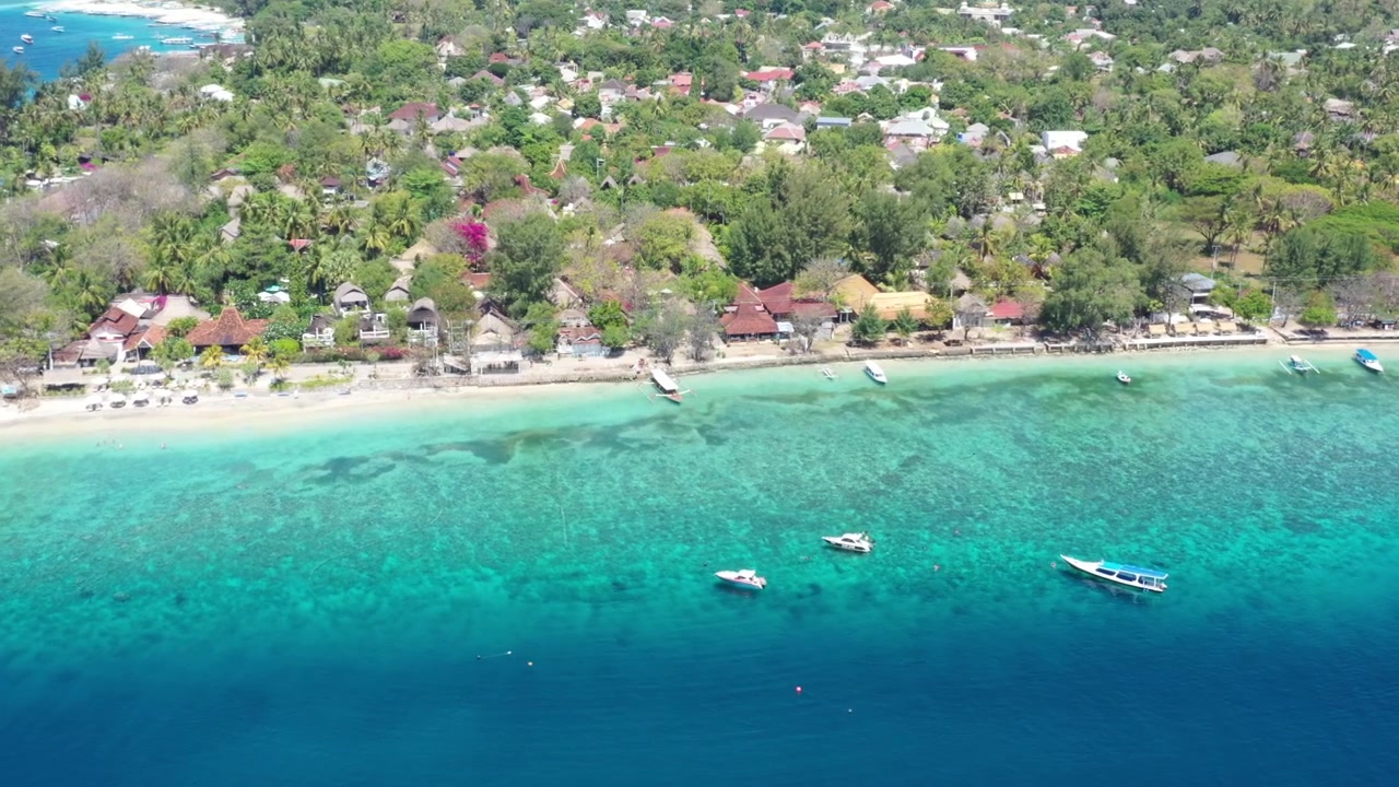 Drone aereo vola verso il basso e si avvicina a Gili Trawangan, rivelando la riva di sabbia bianca, edifici fronte mare, barche, alberi verdi e acqua blu, Lombok, Indonesia. B-roll cinematografico di isola tropicale