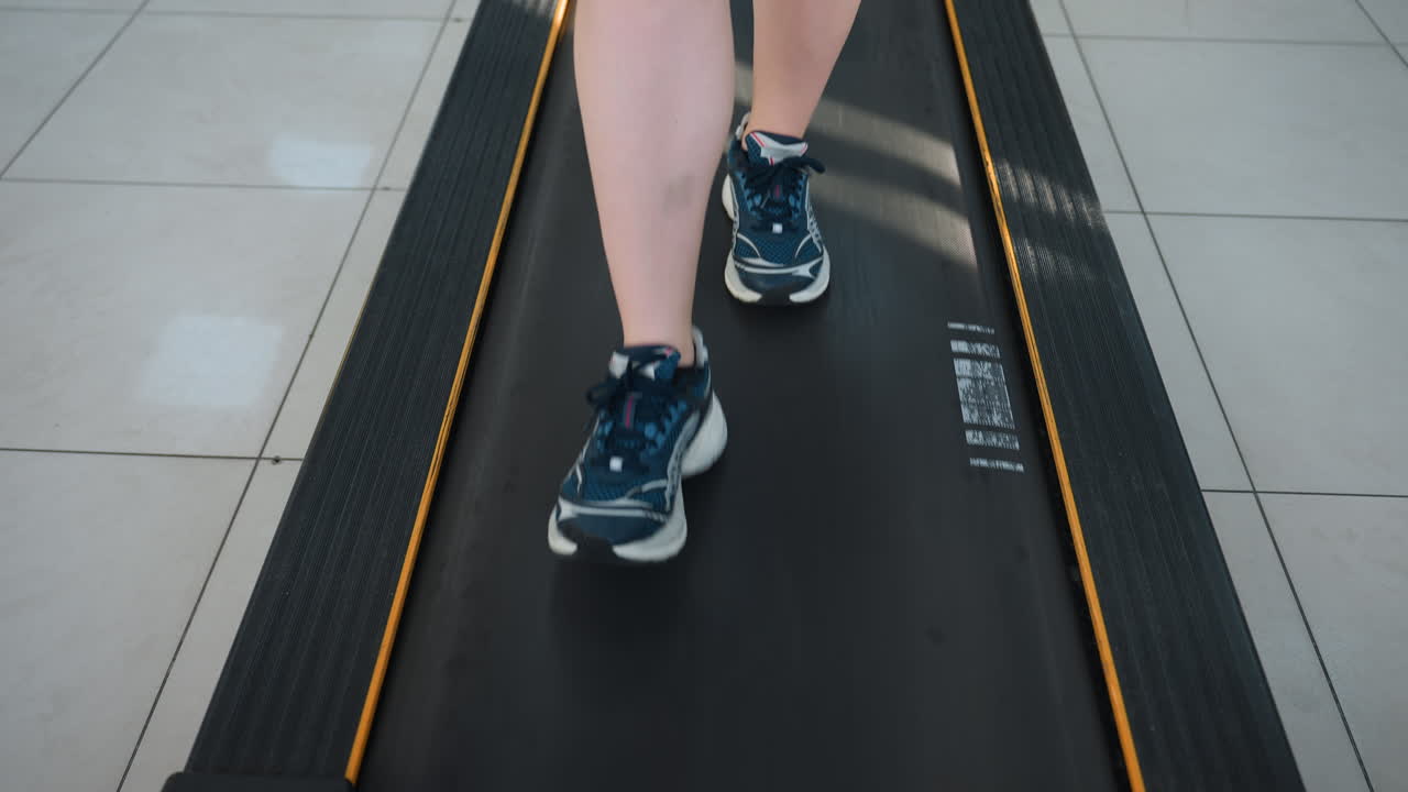 leg view of person walking on treadmill with tiled gym floor, dynamic movement captured from low angle, athletic shoes in motion, sunlight reflection on belt, urban fitness environment
