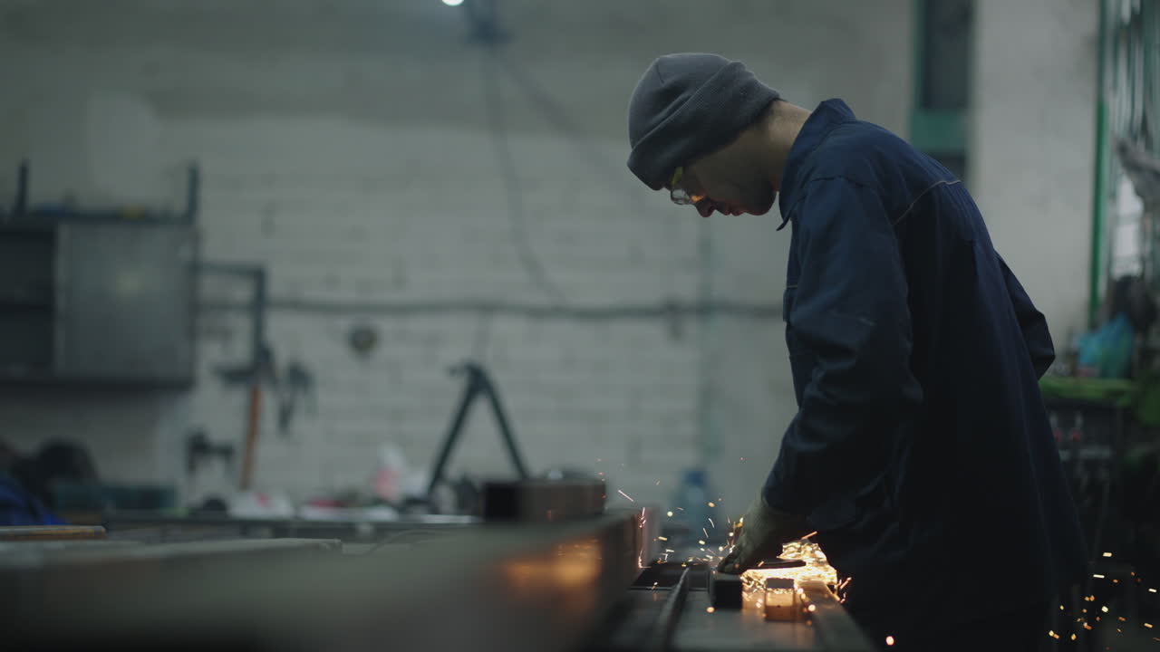 The man works in a factory. A man in protective glasses works with metal grinding polishing and stripping steel metal structures.