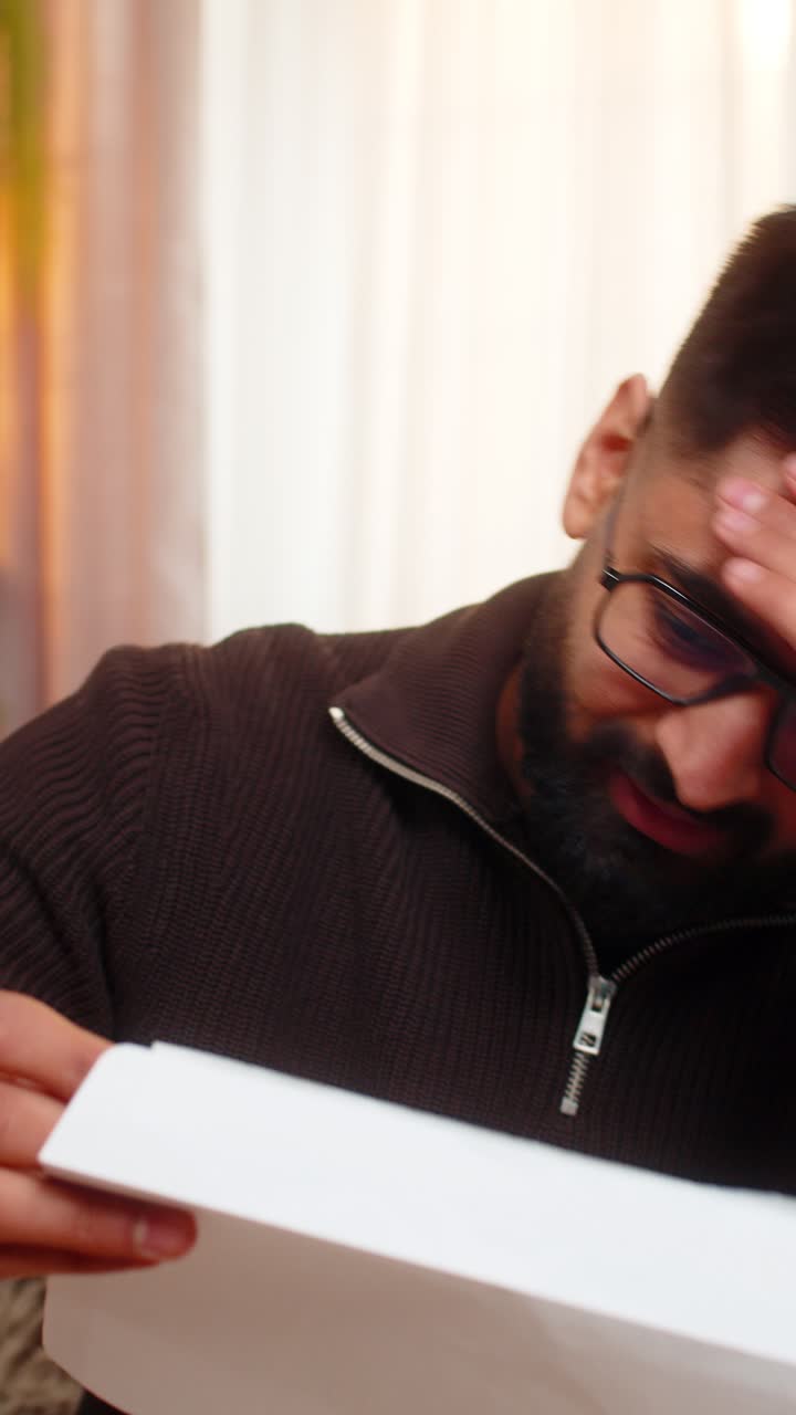 Indian man on sofa opens envelope and looks sad after reading bad news in official rejection letter