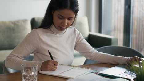 Mujer-De-Raza-Mixta-Trabajando-En-La-Computadora-Y-Tomando-Notas-En-Casa.