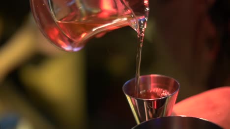 gold colored alcohol being tipped from glass pouring cup into stainless steel shot cup then tipped into stainless steel cocktail mix cup filmed as close up in slow motion