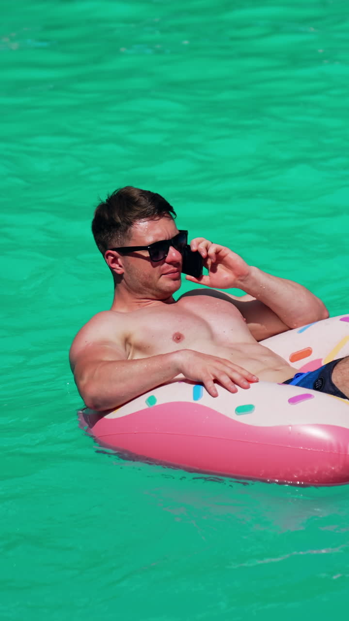 Sunbathing Caucasian man sitting in the inflatable circle talking on the phone. Male wearing sunglasses resting outdoors. Vertical video