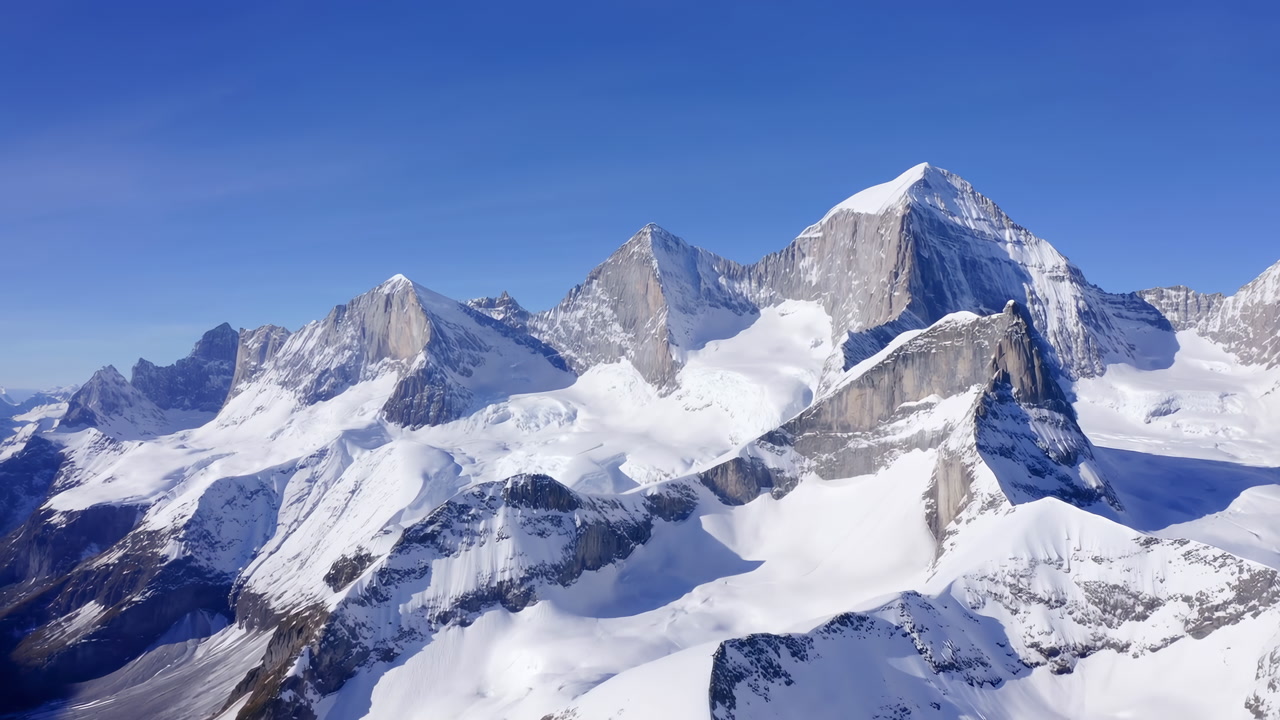 Snow Covered Alpine Peaks Under Clear Blue Sky, Glacial Ridges Bathed In Bright Sunlight, Panoramic Aerial