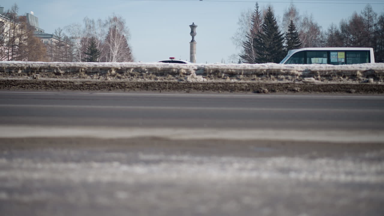 cars moving on both sides of city road in winter, snow piled along median while exhaust drifts, pedestrian shadow stretches across asphalt, trees and power wires frame scene under clear blue sky