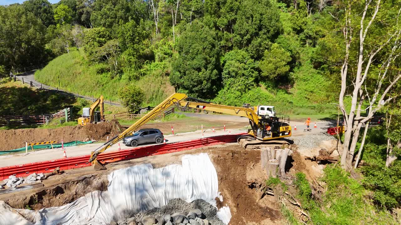 Aerial footage of construction site with excavator and vehicles in lush, green landscape under bright sunlight