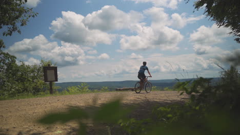 Tiro-Estático-En-Cámara-Lenta-De-Un-Joven-Montando-En-Bicicleta-De-Montaña-En-Un-Sendero-Fuera-De-La-Carretera-Con-Vistas-Al-Cielo-Azul-Brillante-Con-Nubes-Blancas-En-Un-Bosque-En-Pensilvania-En-El-Verano