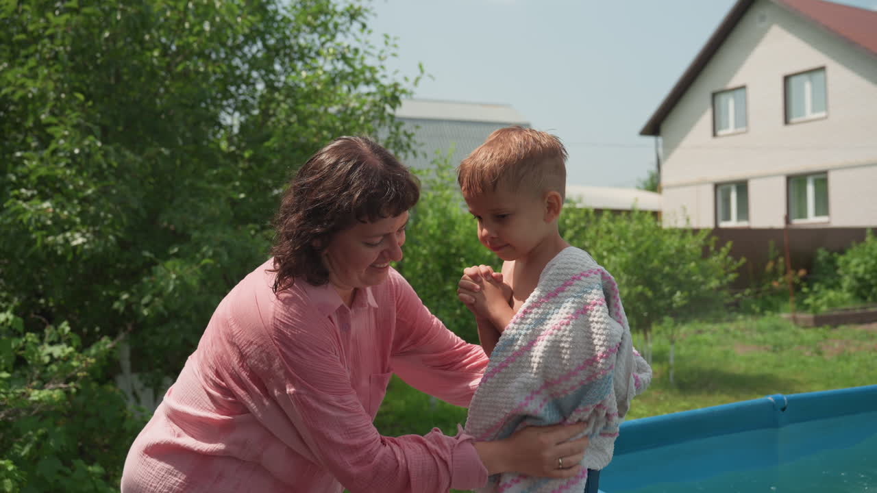 Caucasian Mother Drying Son With Towel By Poolside, Gentle Backyard Care, Warm Summer Light, Suburban House In Background, Affectionate Hug And Playful Splash Marks, Cozy Family Moment