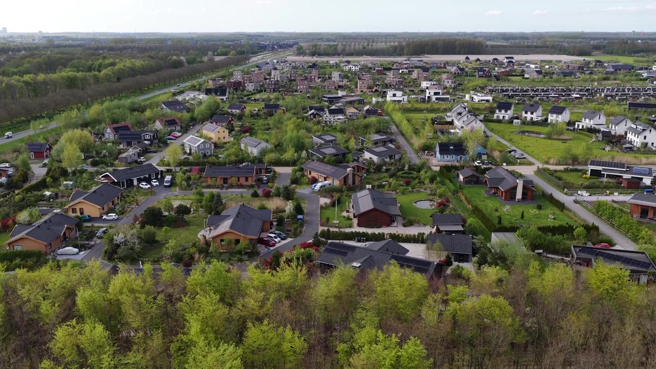 Droneperspektiv, der flyver baglæns og vipper op over en frodig forårsskov med nye grønne blade, der gradvist afslører et moderne forstadskvarter med parcelhuse og grønne haver