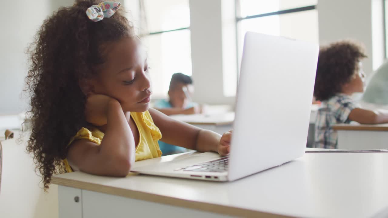 Video of bored african american girl sitting at desk with laptop during lesson in classroom