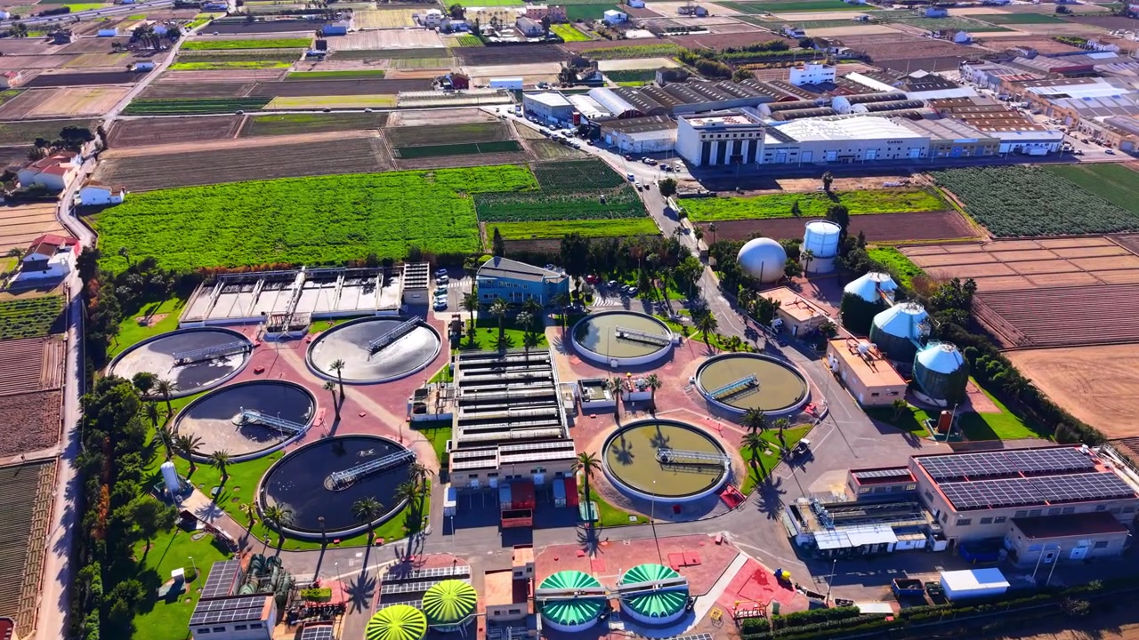 Panoramic aerial view of a large scale industrial purification facility located within a patchwork of agricultural green fields. Vast water treatment plant surrounded by farm land.
