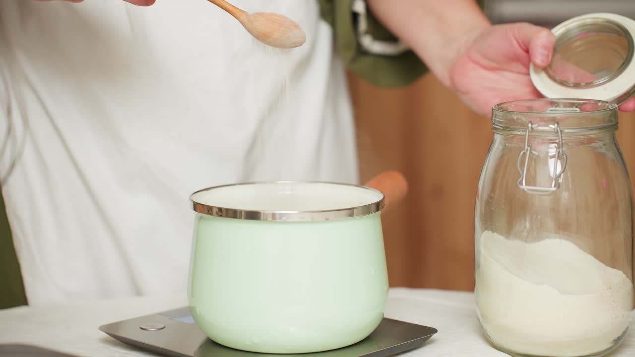 Chef in green outfit and white apron opens sugar jar, scoops sugar with wooden spoon, and gently pours it into mint green pot on scale as fine sugar particles float visibly in air