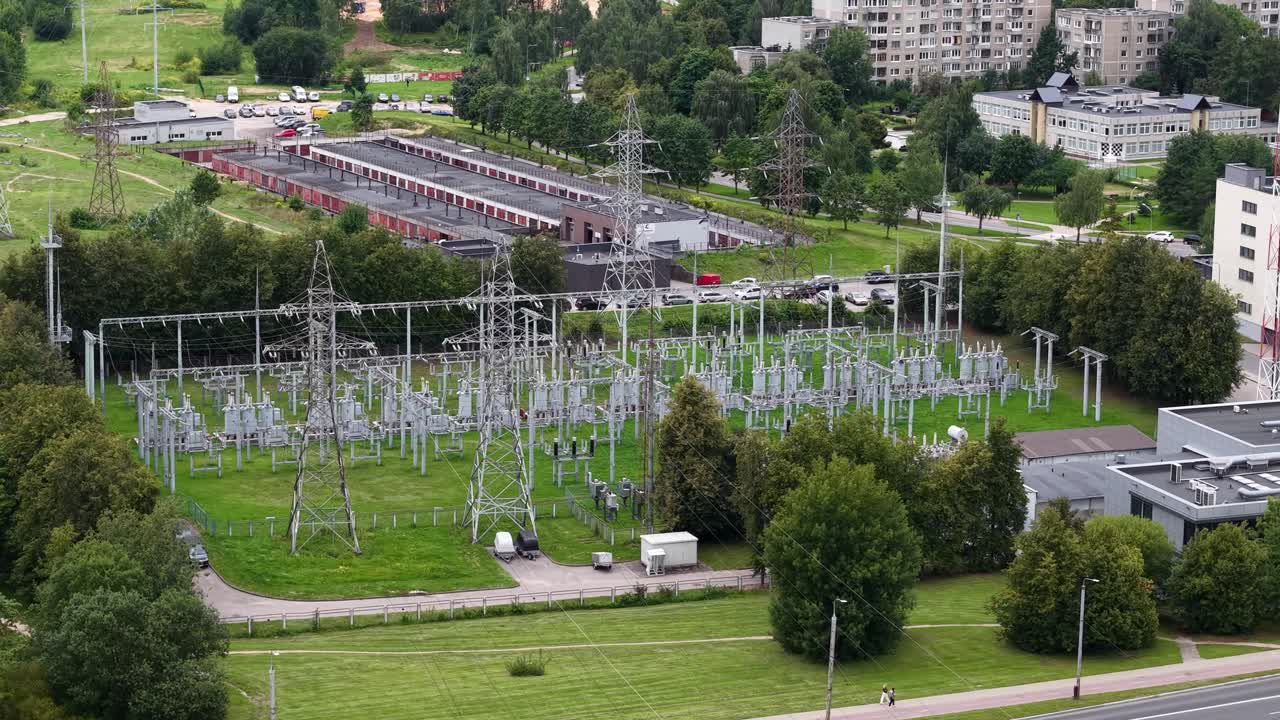 Drone aerial reveals a high-voltage substation and transmission pylons beside suburban buildings and green parkland, showcasing electricity distribution infrastructure