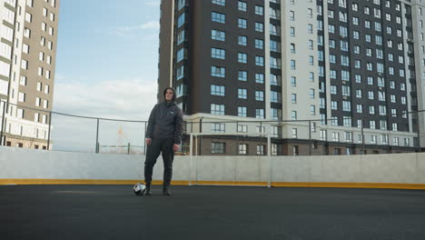 sportsman practicing leg movements with soccer ball in urban sports arena, background features tall modern office buildings and goalpost under clear sky